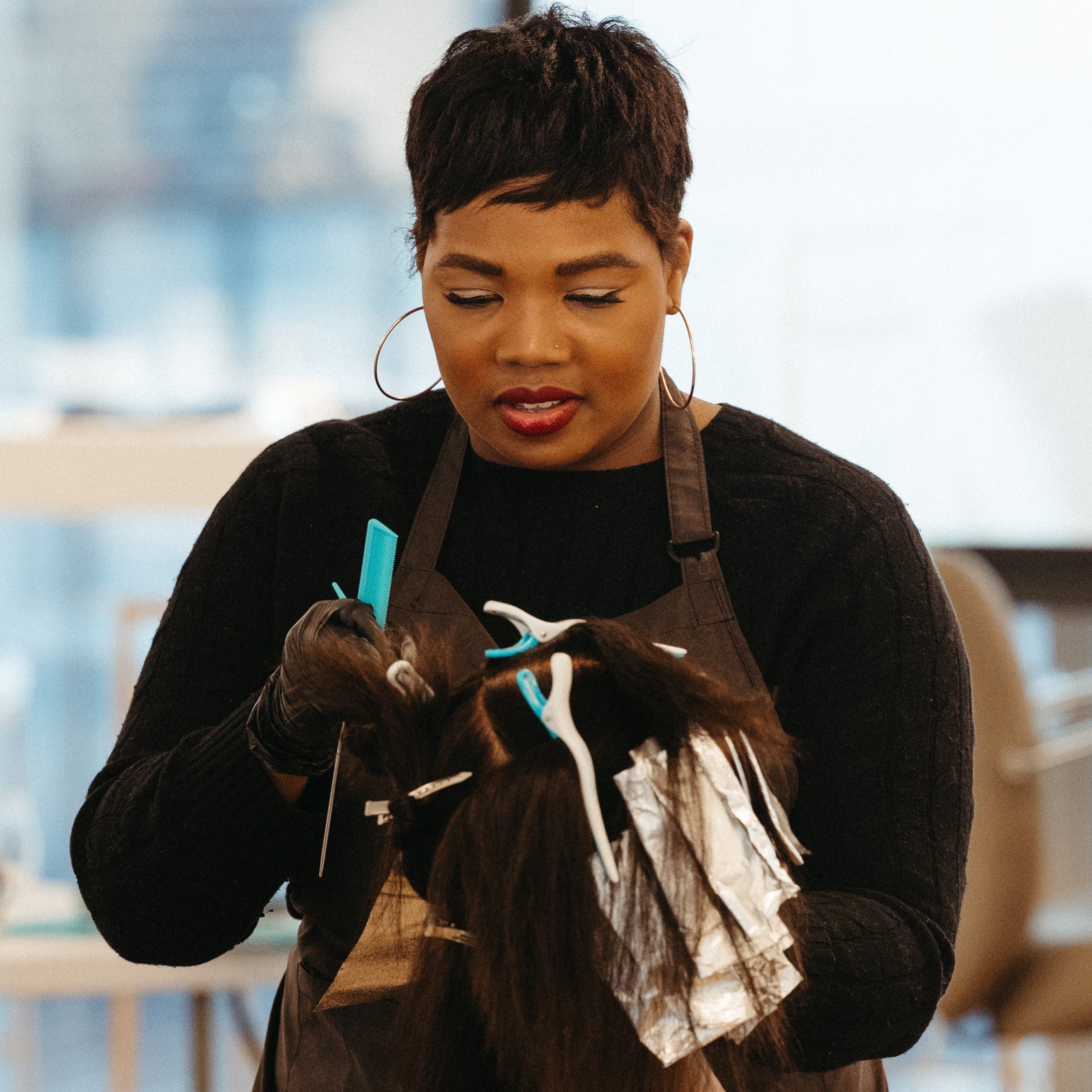 A woman with short dark hair, wearing a black long-sleeve top and a brown hair styling apron, is shown from the chest up in a salon setting. She is holding a hair extension or wig with white adhesive and silver foil, alongside blue and white hair styling tools and bottles.
